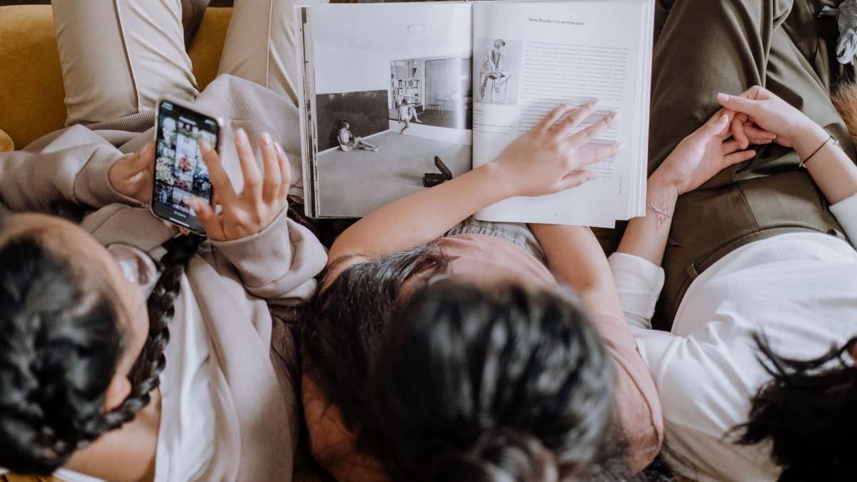 Three young women relaxing on a couch reading and using a smartphone, embodying leisure and togetherness.
