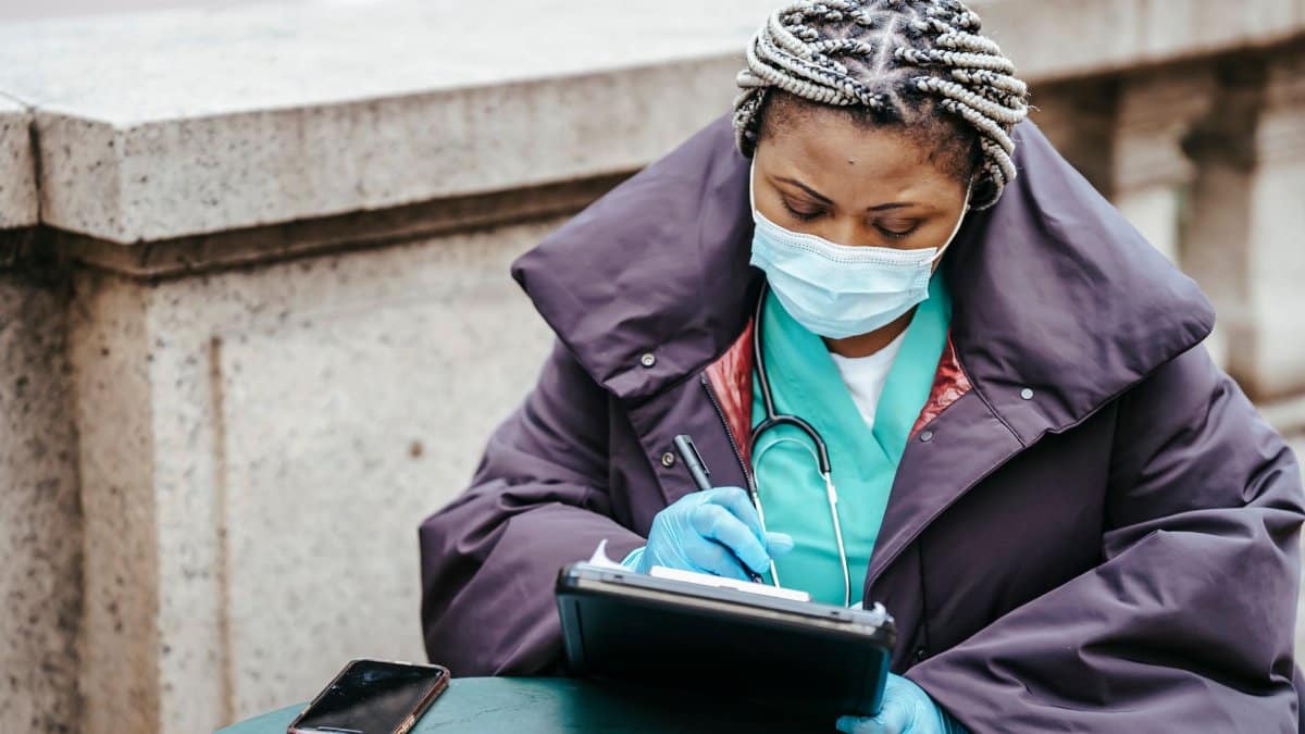 Middle aged African American female medic in outerwear and mask taking notes on paper at table with cellphone in city