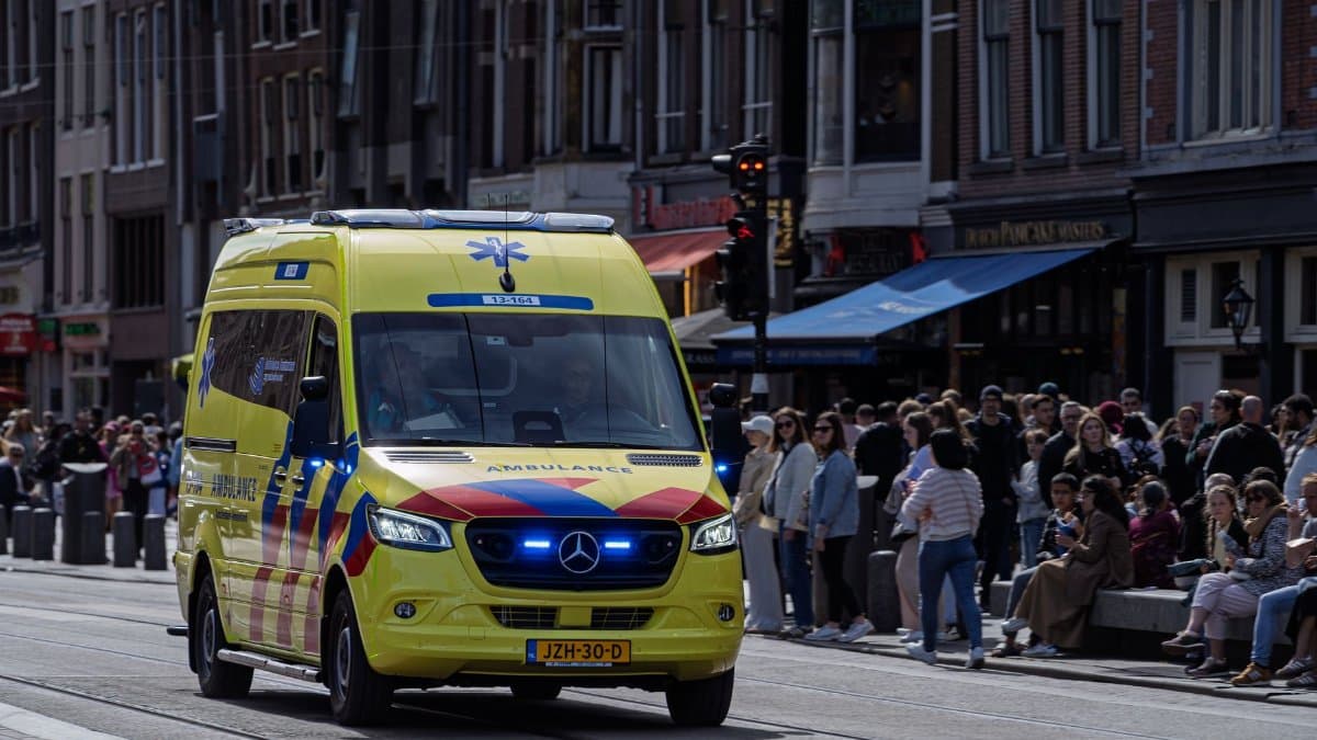 A bright yellow ambulance navigates through a busy European city street with a crowd watching.