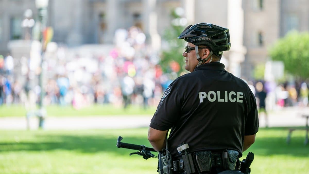 Back view of anonymous police officer in helmet and glasses and summer uniform standing with bicycle in front of protesting crowd