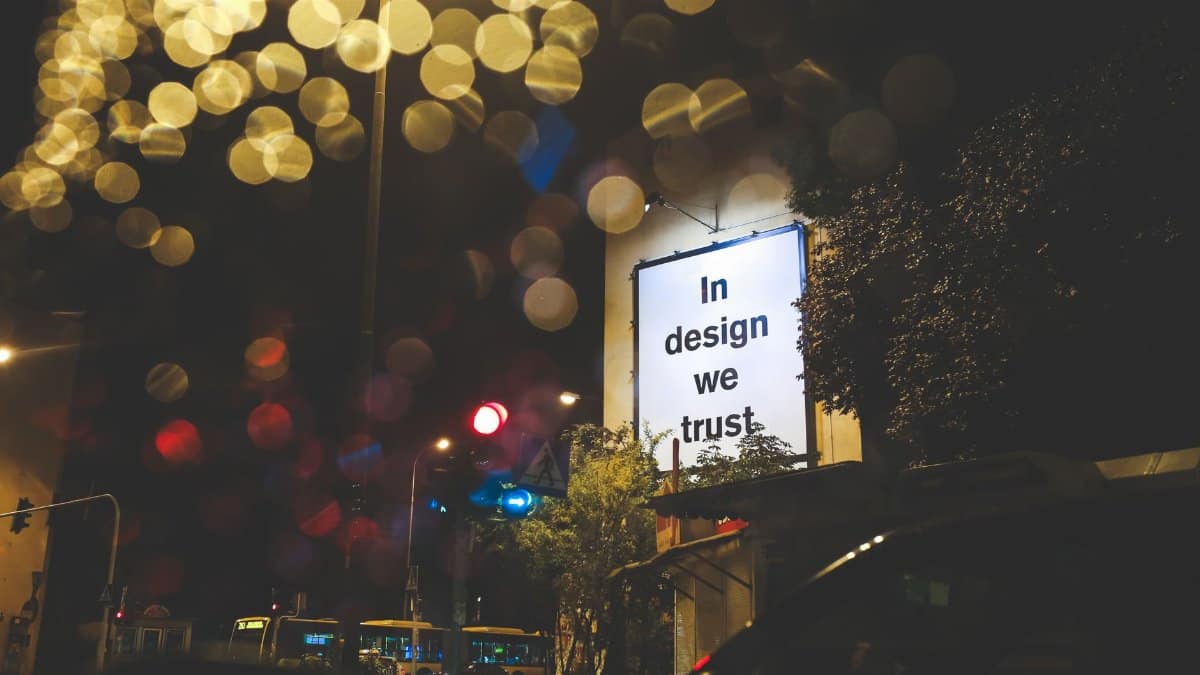 A city street at night featuring an illuminated billboard with the phrase 'In design we trust.'