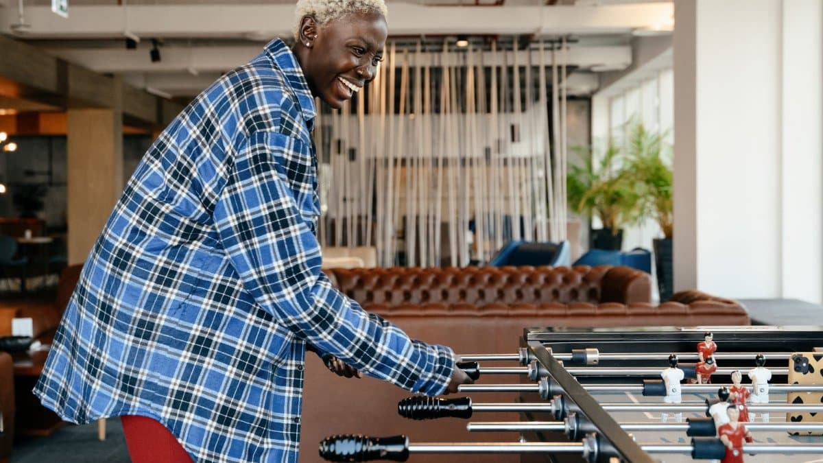 Side view of smiling African American female in casual clothes standing and playing foosball in living room at home