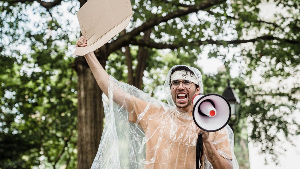Man holding megaphone and cardboard at protest rally in park.