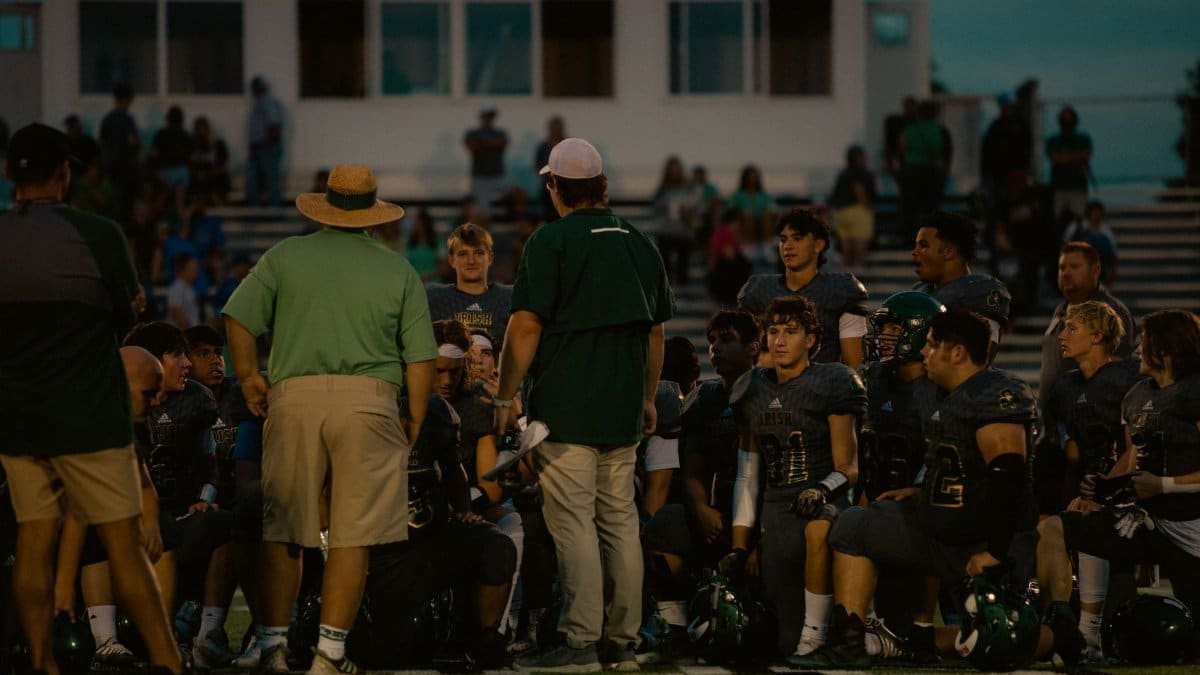 Football team huddles with coaches during a game at a stadium under the evening sky.