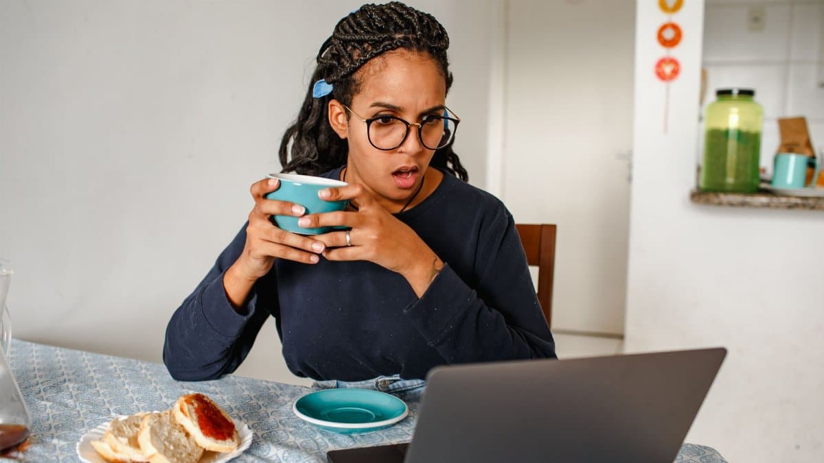 Young woman with glasses looks surprised while drinking coffee and using a laptop indoors.