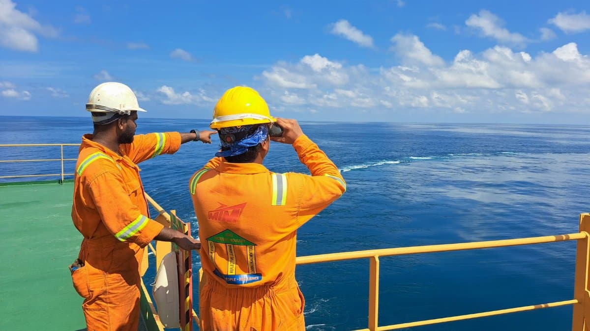Two workers on an oil rig deck wearing safety gear inspect the ocean horizon under a clear sky.
