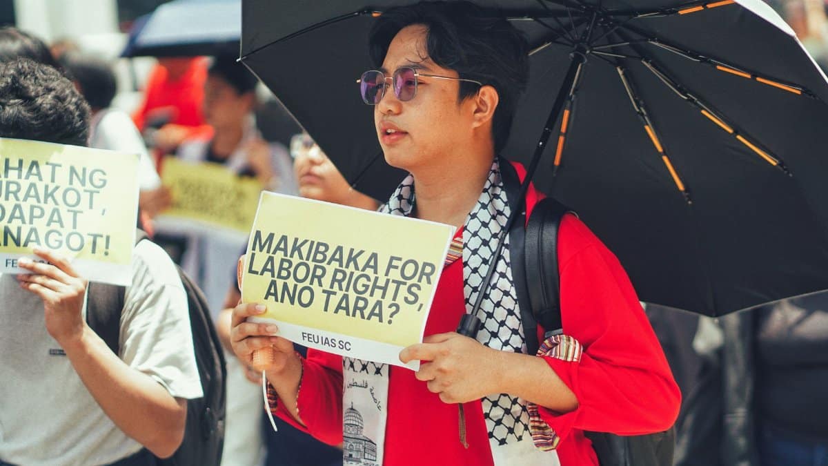 Protest for labor rights featuring diverse participants holding placards and an umbrella for shade.