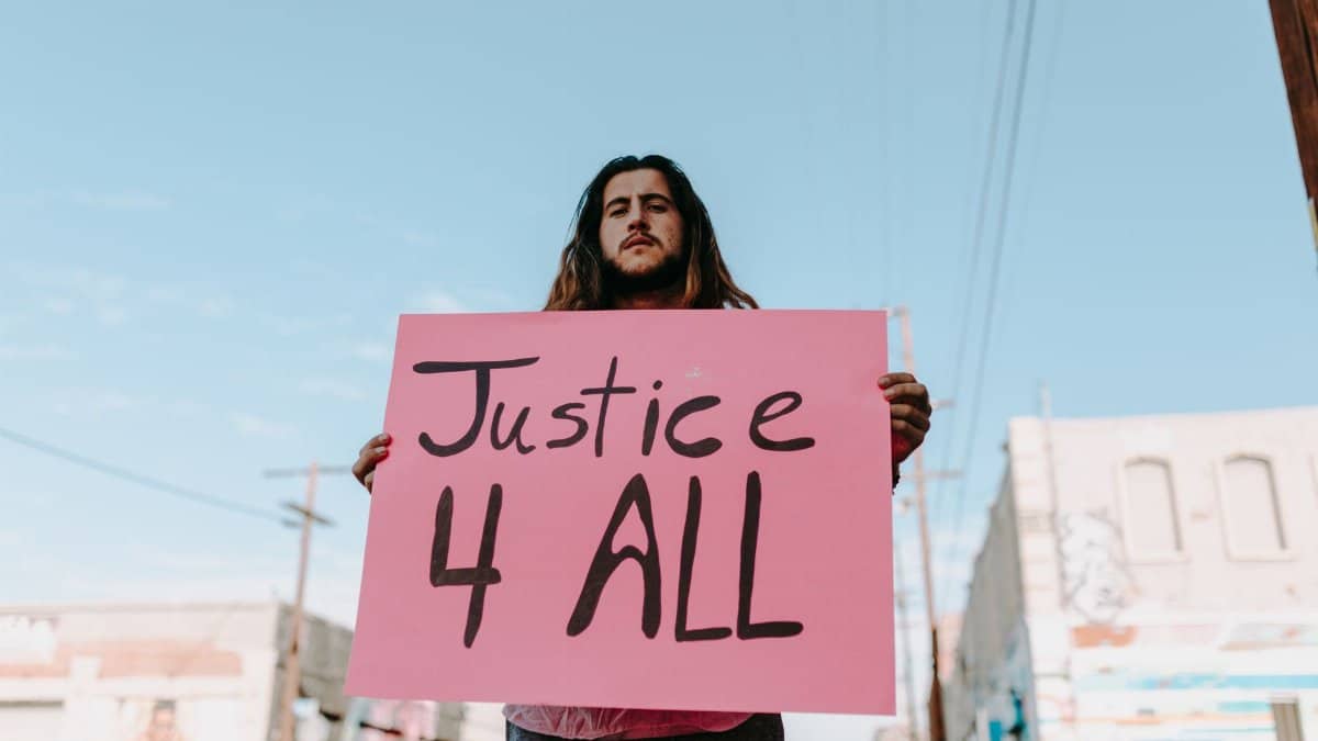 Man holding a pink sign reading 'Justice 4 All' at a peaceful protest in an urban setting.