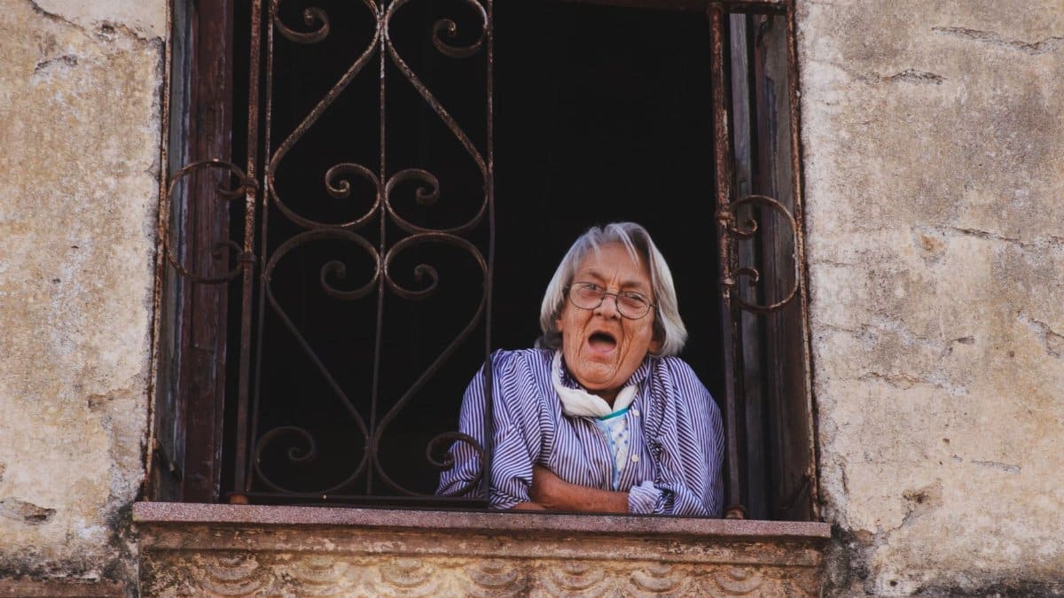 A senior woman peers out of a rustic window with decorative iron bars on a stone building.