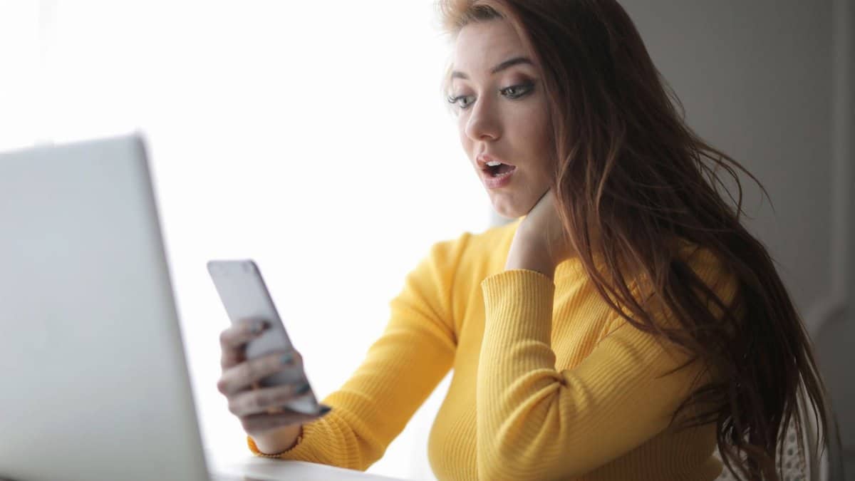 Young woman in yellow sweater looks surprised at phone while working on a laptop indoors.