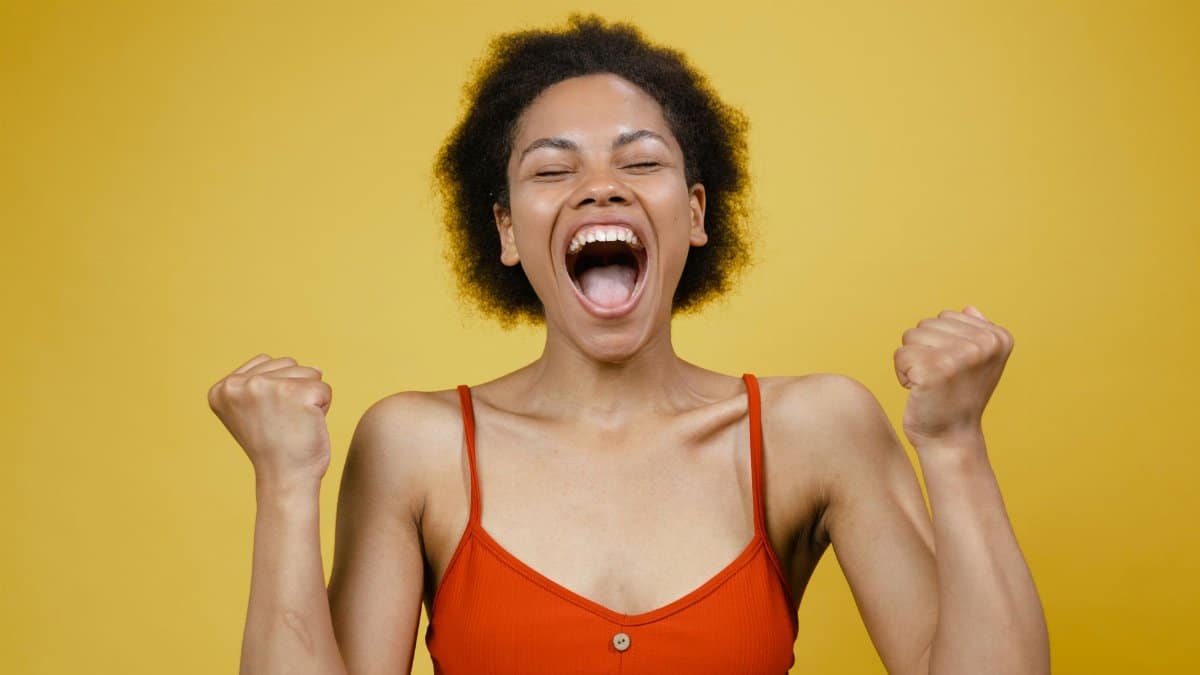 A cheerful woman in a red top expressing excitement and happiness against a vibrant yellow background.