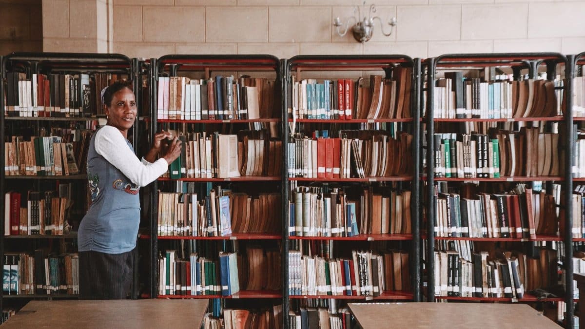 A woman stands beside tall bookshelves filled with books in a library setting.