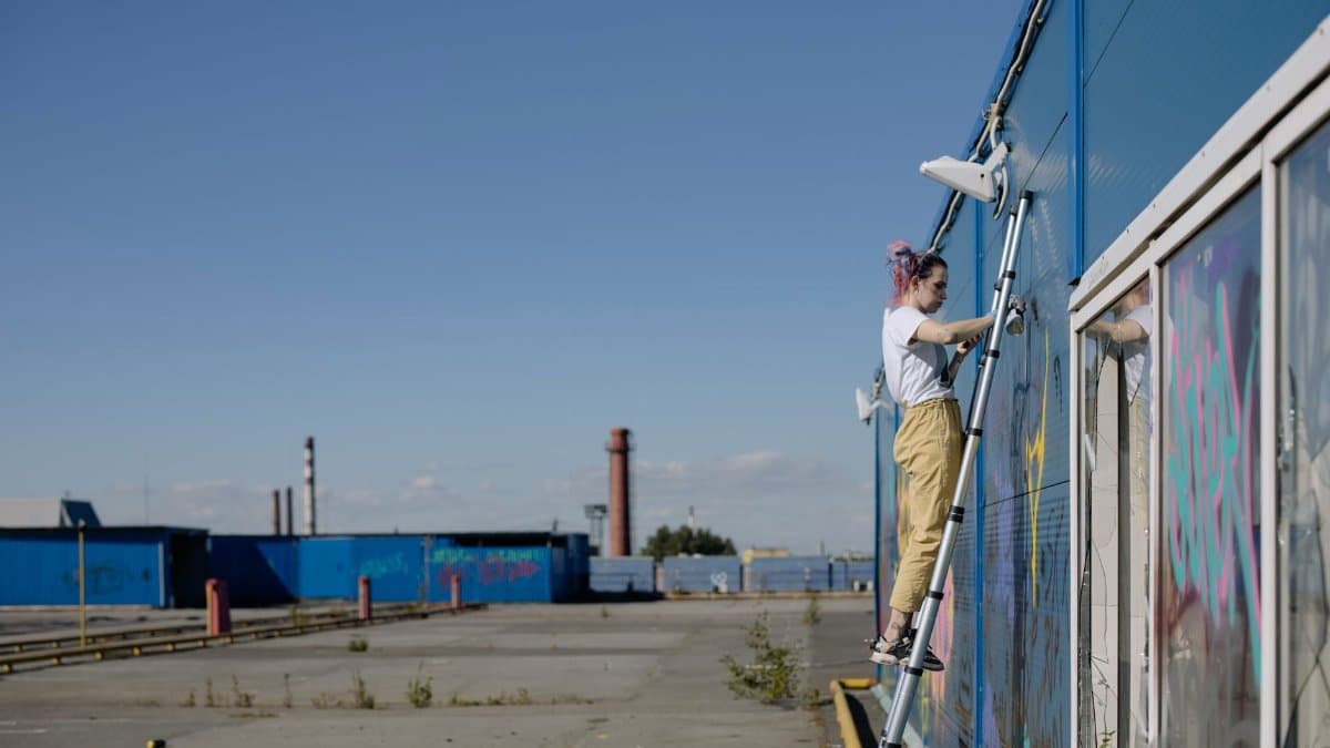 A woman stands on a ladder painting graffiti on an industrial building in daylight.