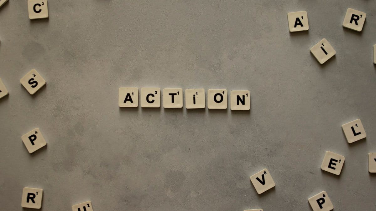 Flat lay of Scrabble tiles spelling 'Action' on a gray background, surrounded by random letters.