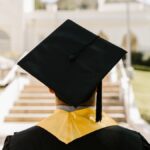 Back view of a graduate in a cap and gown standing outdoors facing a building.
