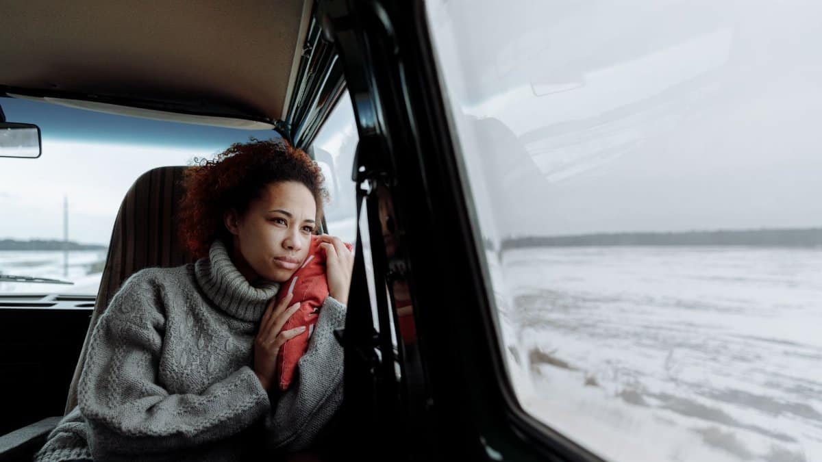 Woman in gray sweater looks out van window at snowy landscape during a winter road trip.
