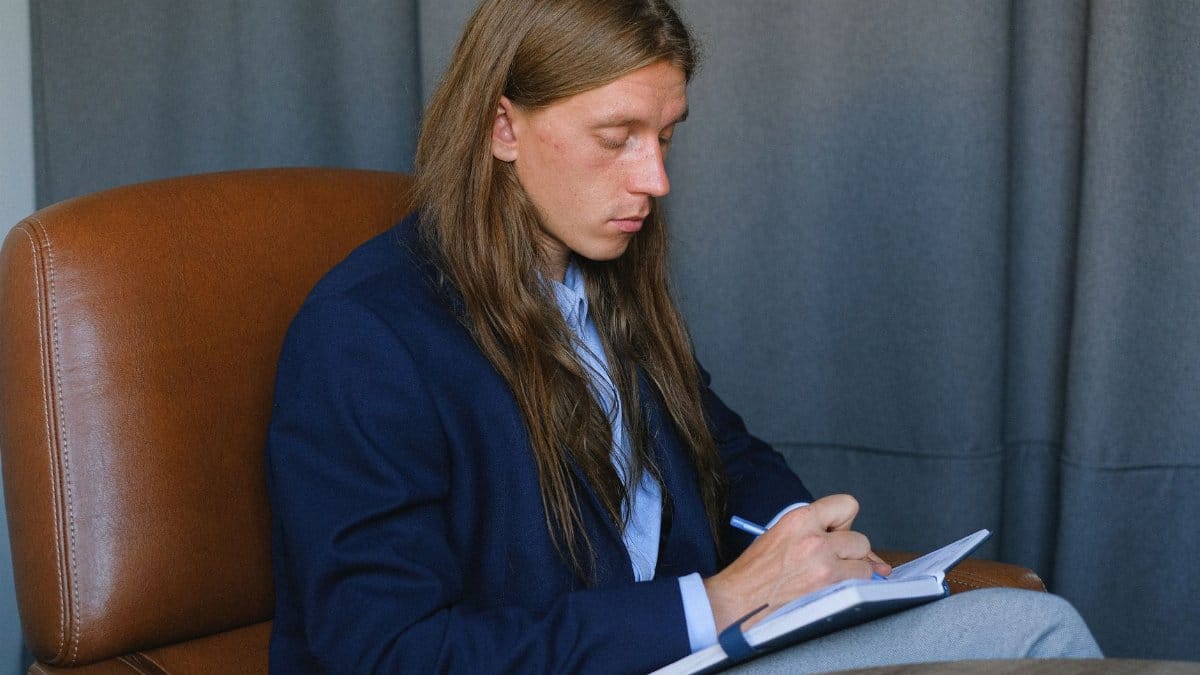 Side view of thoughtful journalist with long hair in stylish formal clothes taking notes in notebook while sitting at table in room against gray curtains