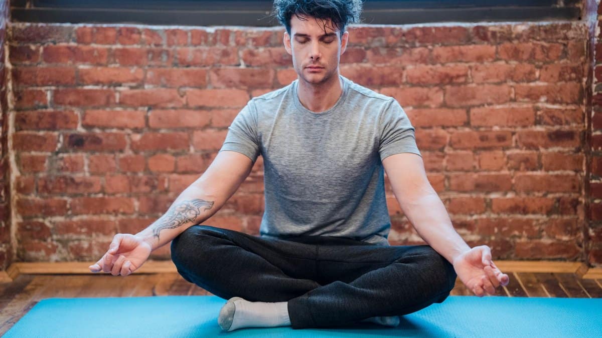 Full body of focused male with closed eyes doing Sukhasana while meditating on yoga mat against brick wall