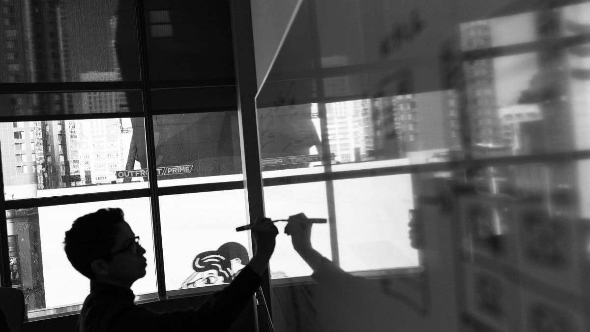 Silhouette of a person writing on a glass board in an office setting, reflecting urban surroundings.