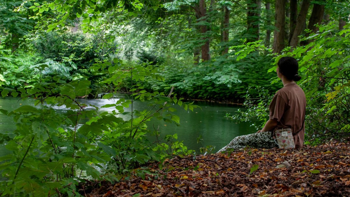 A woman sits on a riverbank surrounded by vibrant green trees, reflecting peacefully.