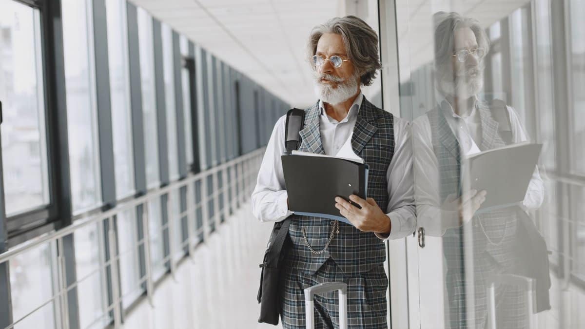 Elderly bearded man in a gray suit holding a folder in a modern glass hallway.