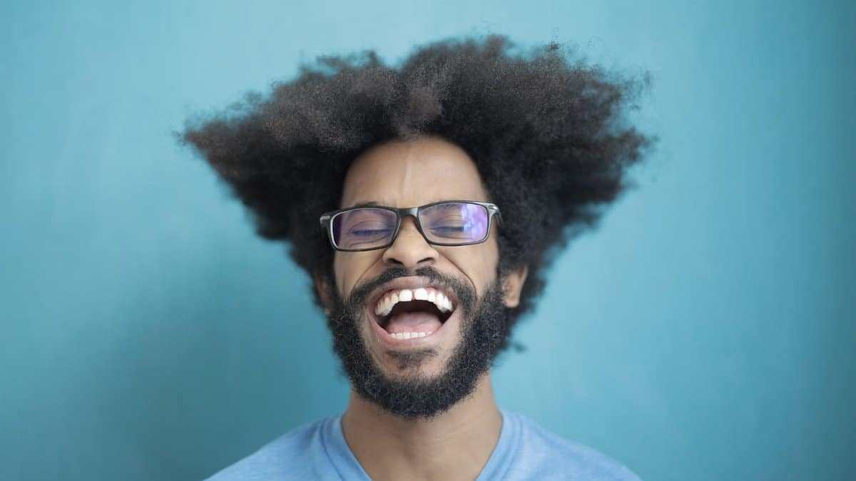Happy man with afro hair and glasses laughing joyfully against a blue background.