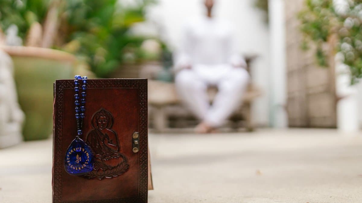 A leather notebook with a Buddhist amulet in focus, symbolizing spirituality, with a person meditating in the background.