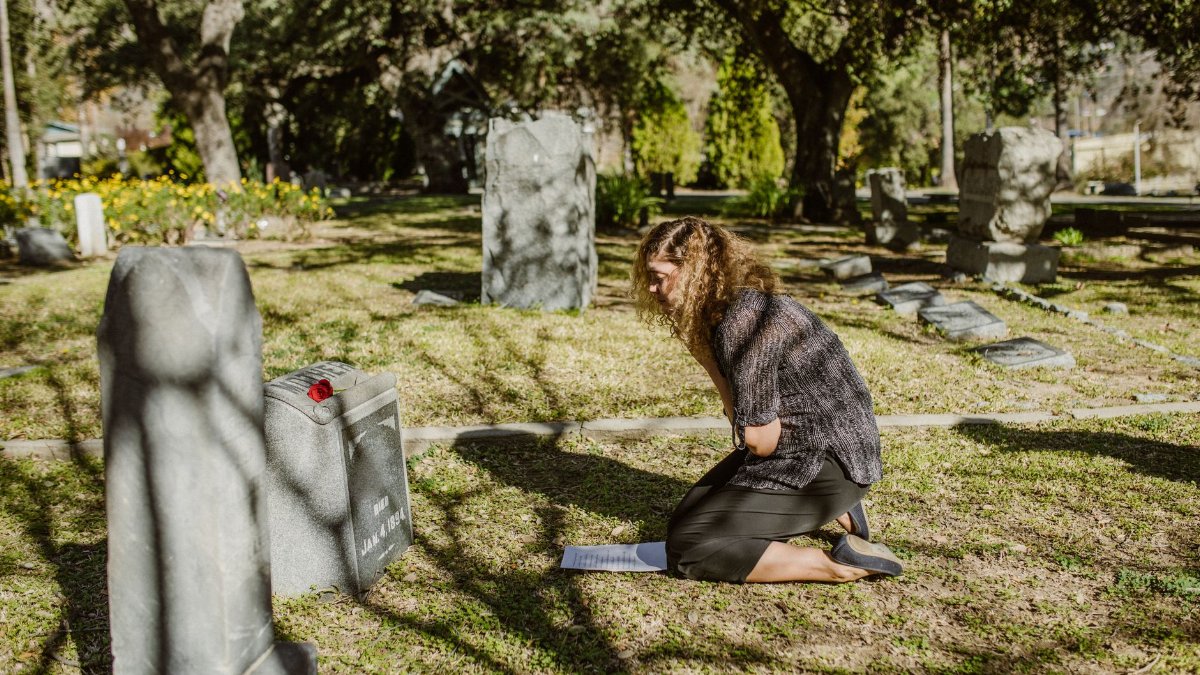 A woman kneels by a gravestone, expressing grief and loss in a sunlit cemetery.