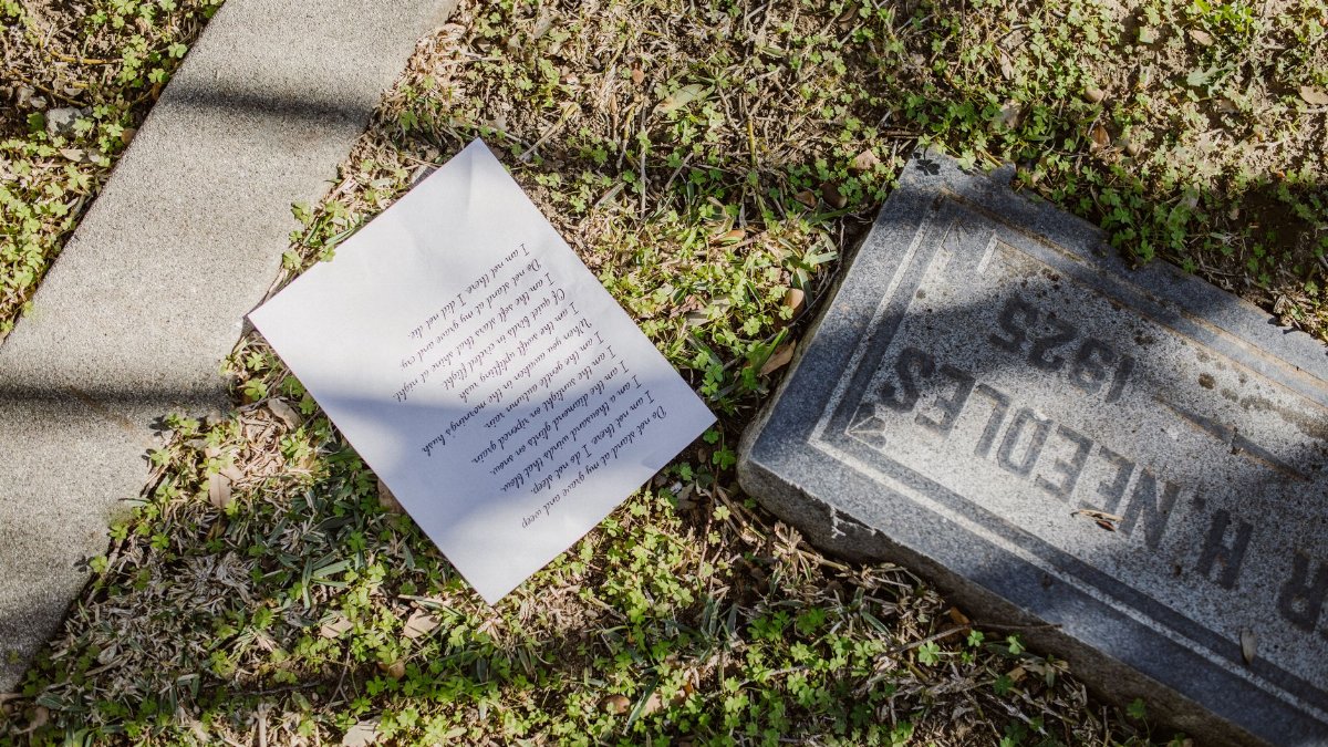 A gravestone and a partially visible letter on grass in a sunlit cemetery.