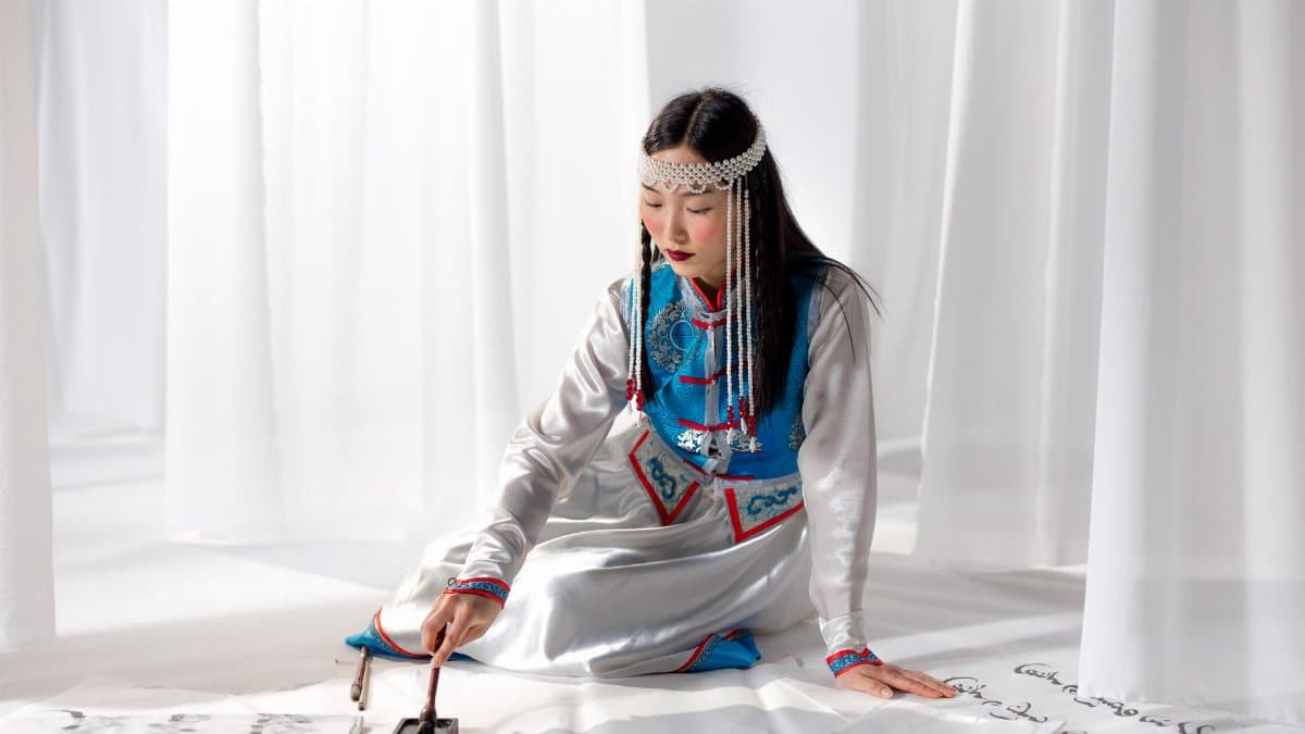 Asian woman practicing traditional calligraphy with tools, wearing cultural attire.