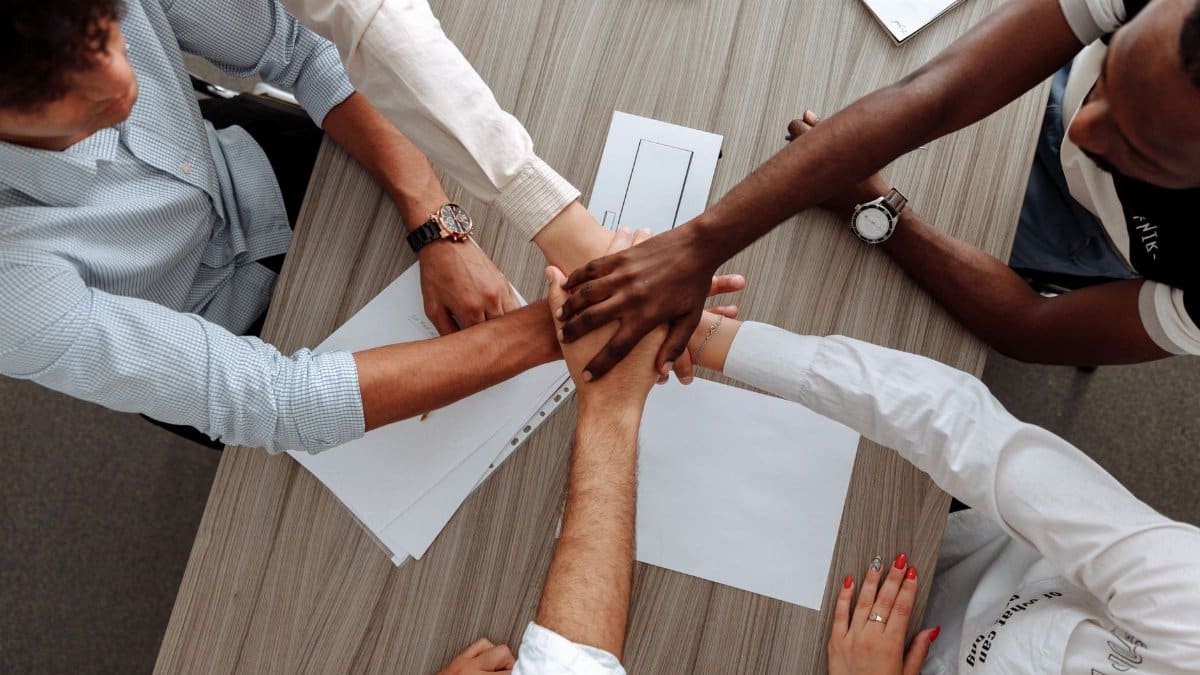 A diverse group of adults join hands in an office, symbolizing teamwork and cooperation.
