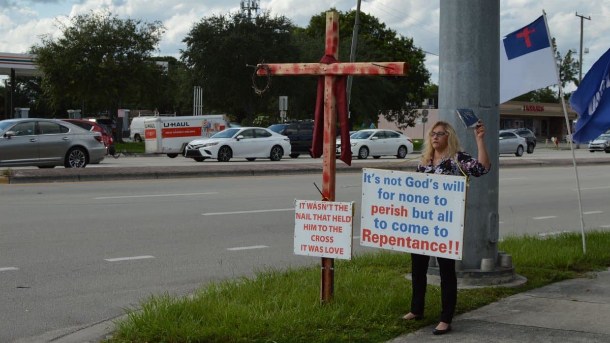 Woman holds religious sign on roadside advocating repentance with cross nearby.