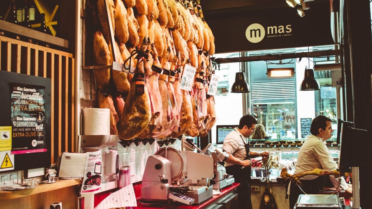 A bustling Spanish ham shop with hanging cured meats and two men serving customers.