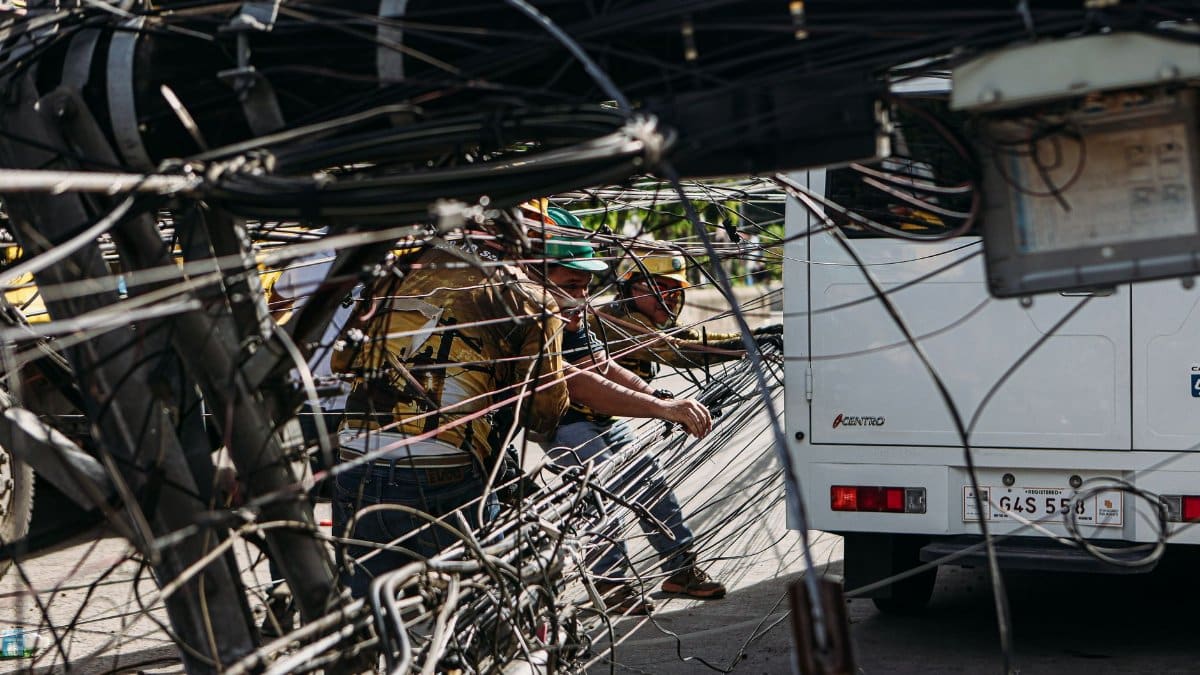 Workers managing tangled power lines by a street setup near a vehicle, highlighting infrastructure challenges.