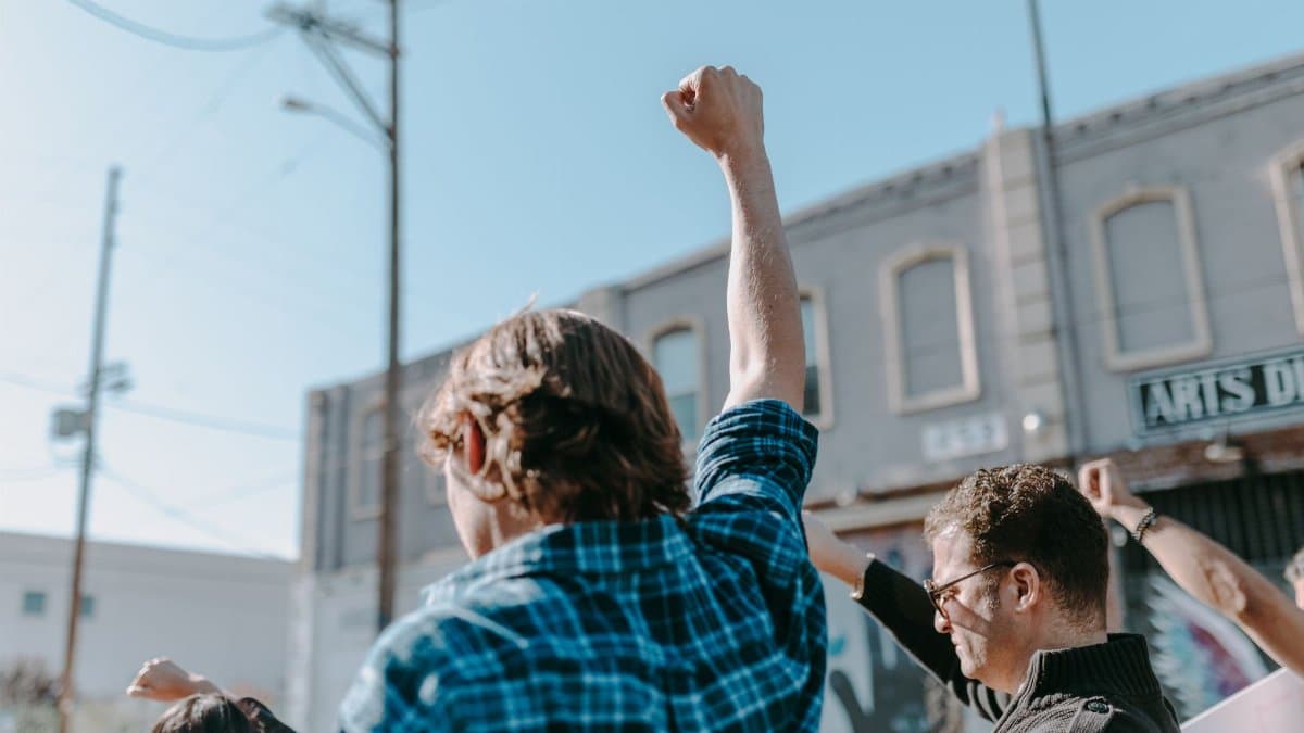 A group of individuals seen from behind raising their fists during a daytime protest outdoors.