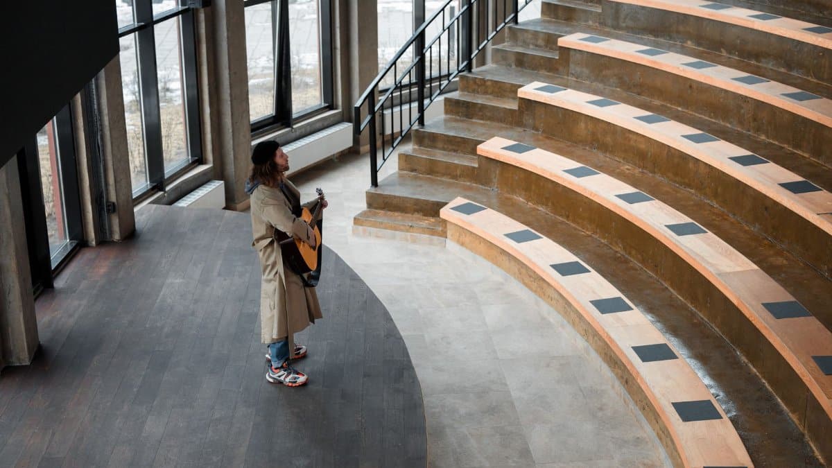 Man playing guitar in a contemporary indoor amphitheater with wooden steps.