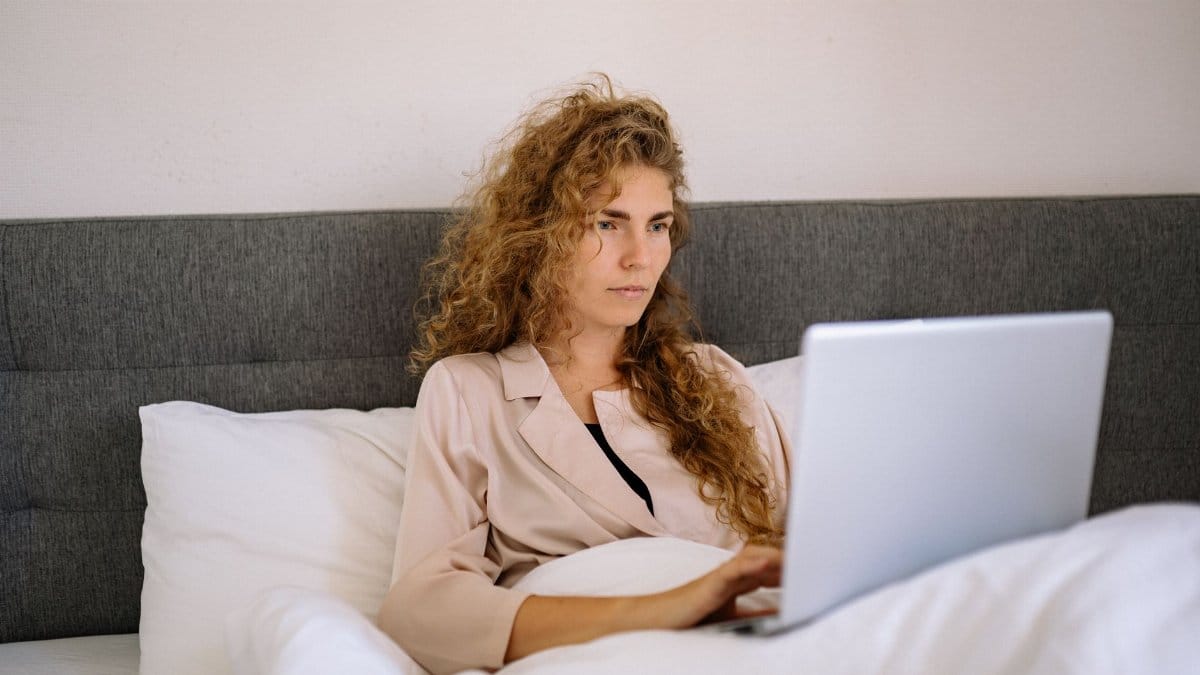 Focused woman with curly hair working on a laptop while sitting in bed in a cozy home setting.