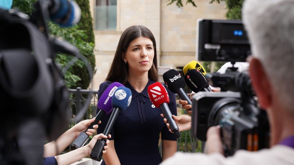 A spokesperson engages with media during an outdoor press interview surrounded by microphones.
