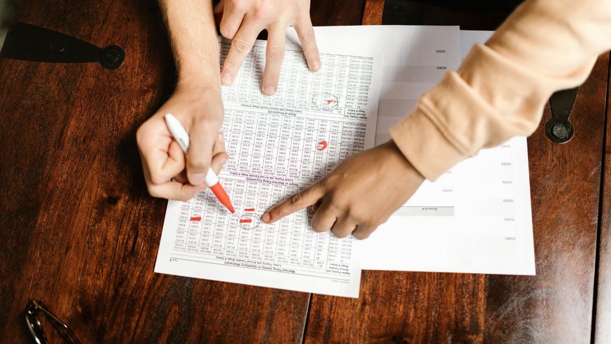 Two hands reviewing and marking financial documents with a red pen on a wooden table, top view.