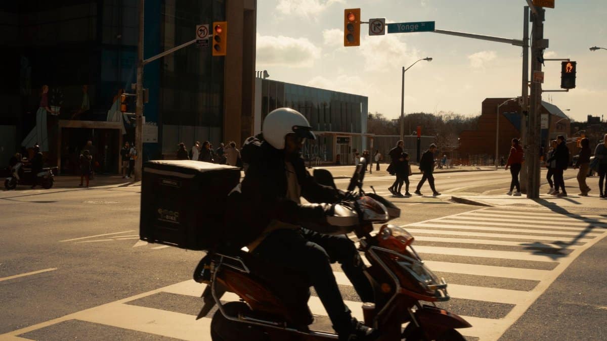 A scooter rider navigates a busy city intersection with pedestrians crossing on a sunny day.