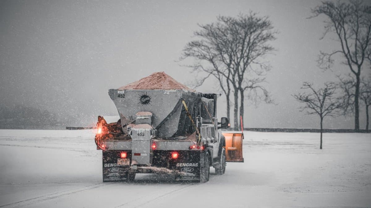 A snowplow truck spreading salt on a snowy road during a winter storm, with bare trees in the background.