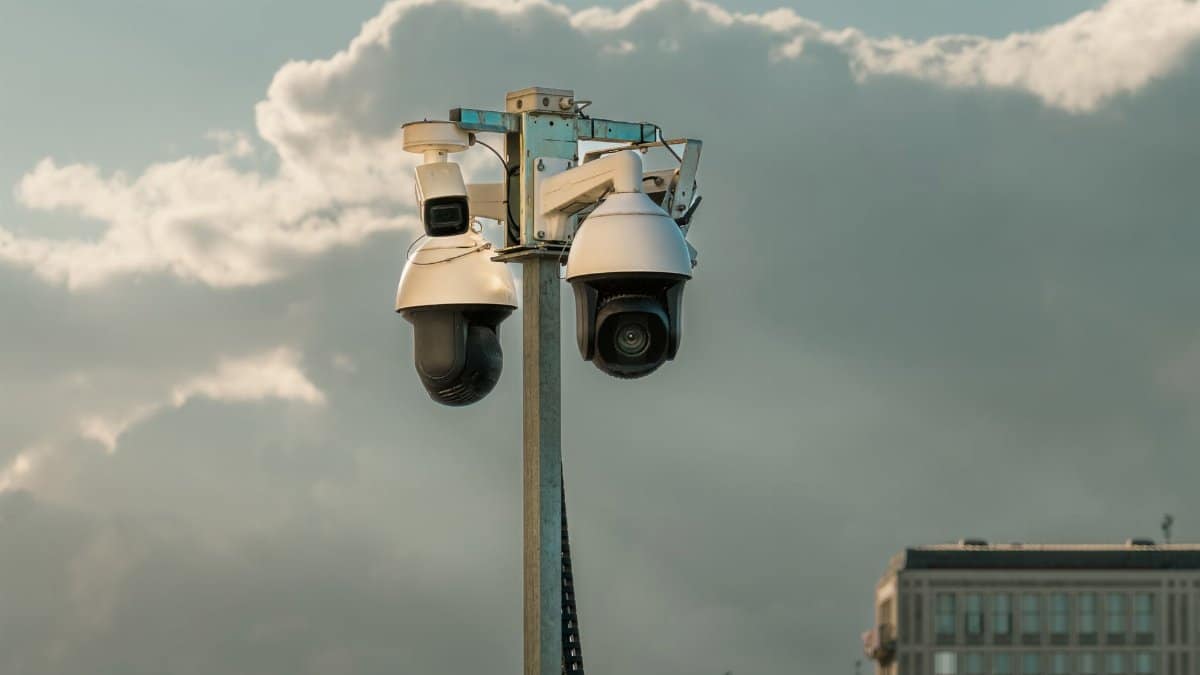 Two modern surveillance cameras mounted on a pole in an urban setting under a cloudy sky.