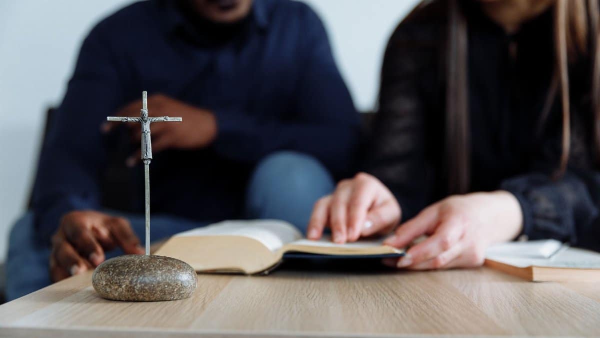 Two individuals study the Bible with a cross on the table, symbolizing faith and devotion.