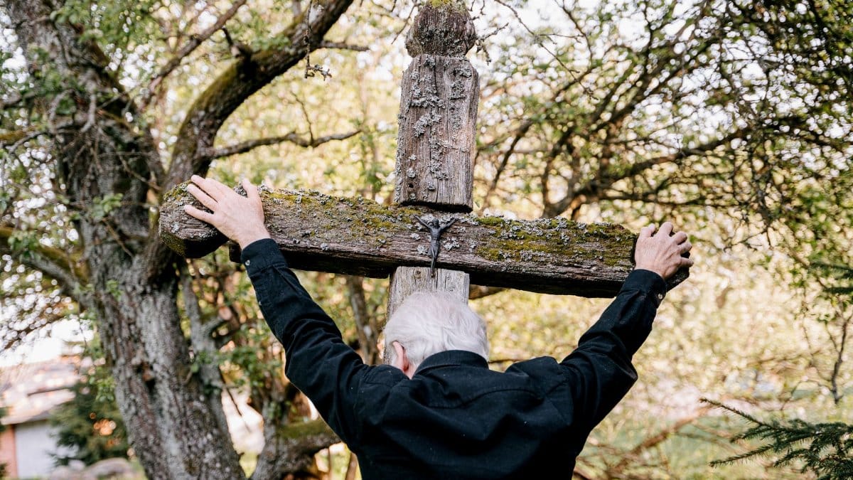 An elderly man embraces a wooden cross, expressing grief and religion in an outdoor setting.