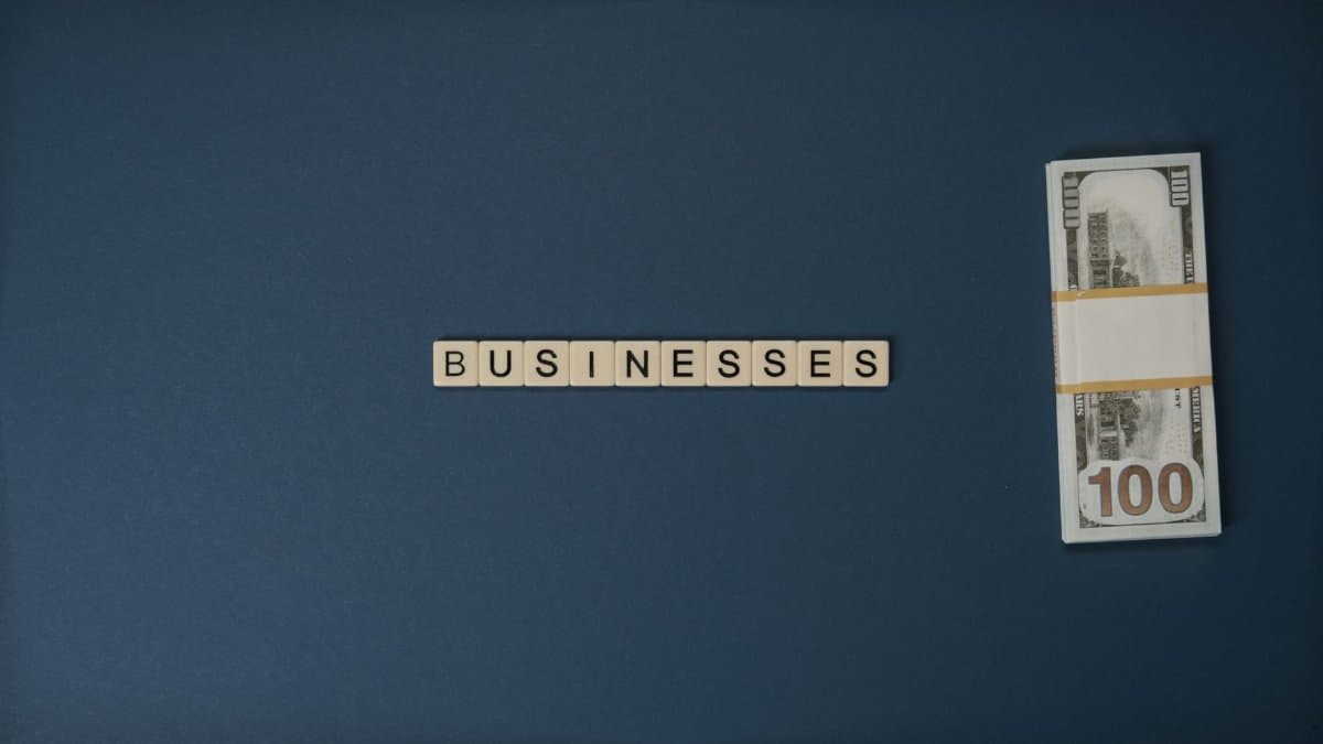 Dollar bills and 'BUSINESSES' spelled with tiles on a blue background, symbolizing finance.