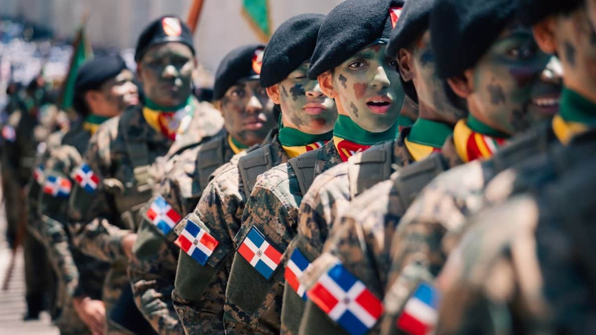 Soldiers in camouflage with face paint in a formation display national flags during a military parade.