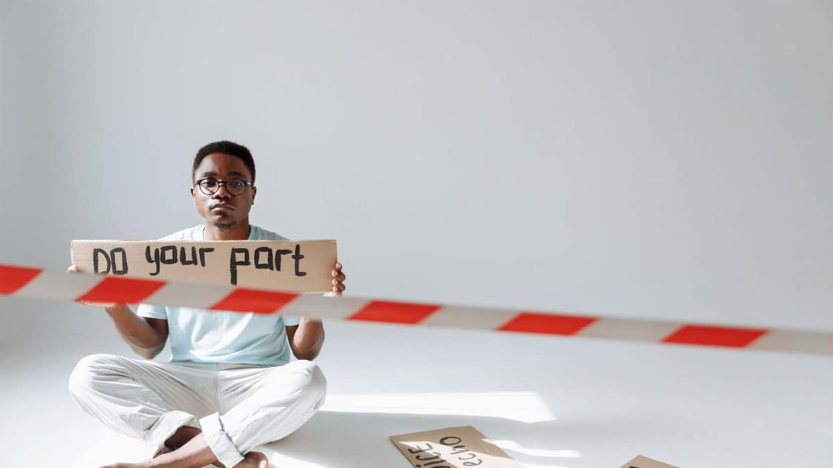 A man sits indoors holding a 'Do Your Part' protest sign with caution tape visible.