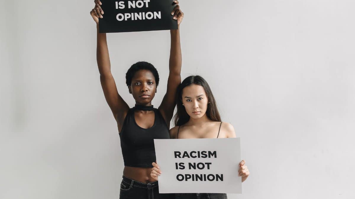 Two women holding 'Racism is Not Opinion' signs on a white background promoting anti-racism awareness.