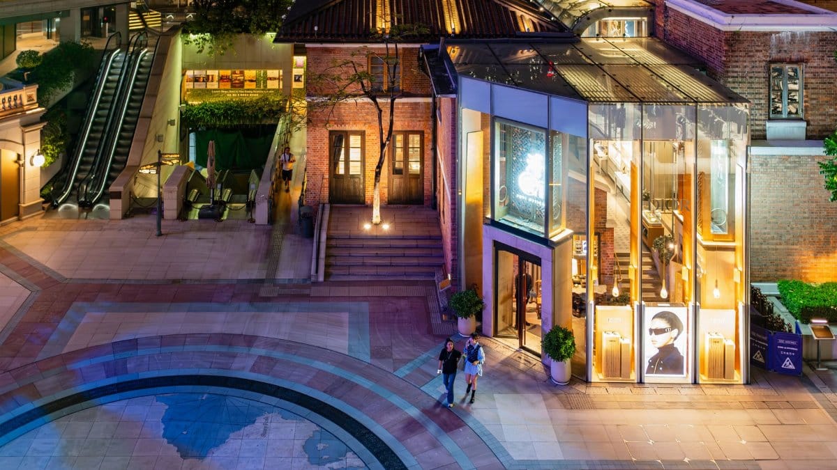 Aerial view of a modern shopping complex with illuminated storefront at night, featuring two people walking on the plaza.