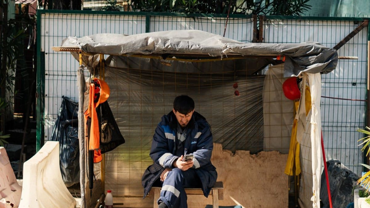 A construction worker sits in a makeshift shelter on a city street, using a phone during a sunny day.